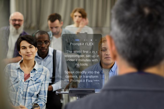 The back of the head of a presenter reading from a teleprompter autocue which shows text on the beamsplitter glass. sitting audience is watching the presenter.