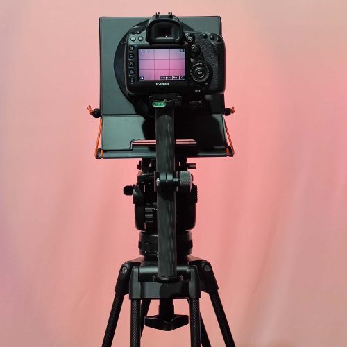 Rear view of Prompt-it MAXI teleprompter mounted on tripod with DSLR camera attached, showing camera screen, tablet platform, hood and support system against a pink background.